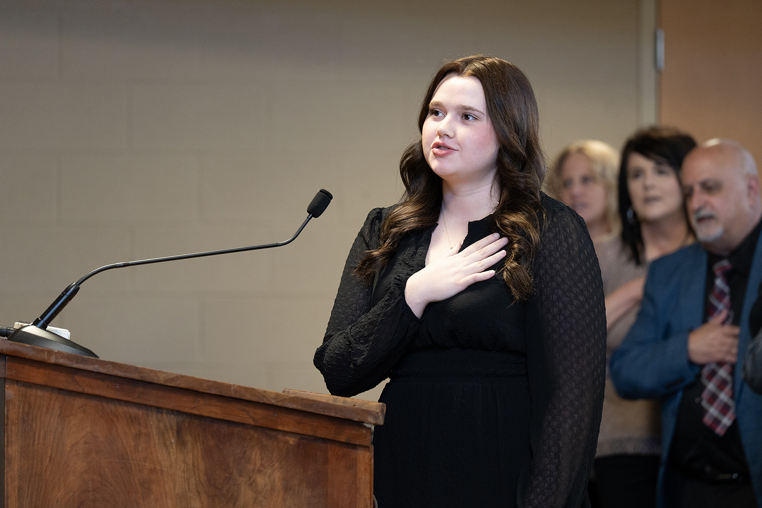 A teenage girl with long, dark hair wearing a black dress stands at a wooden podium with her right hand over her heart.