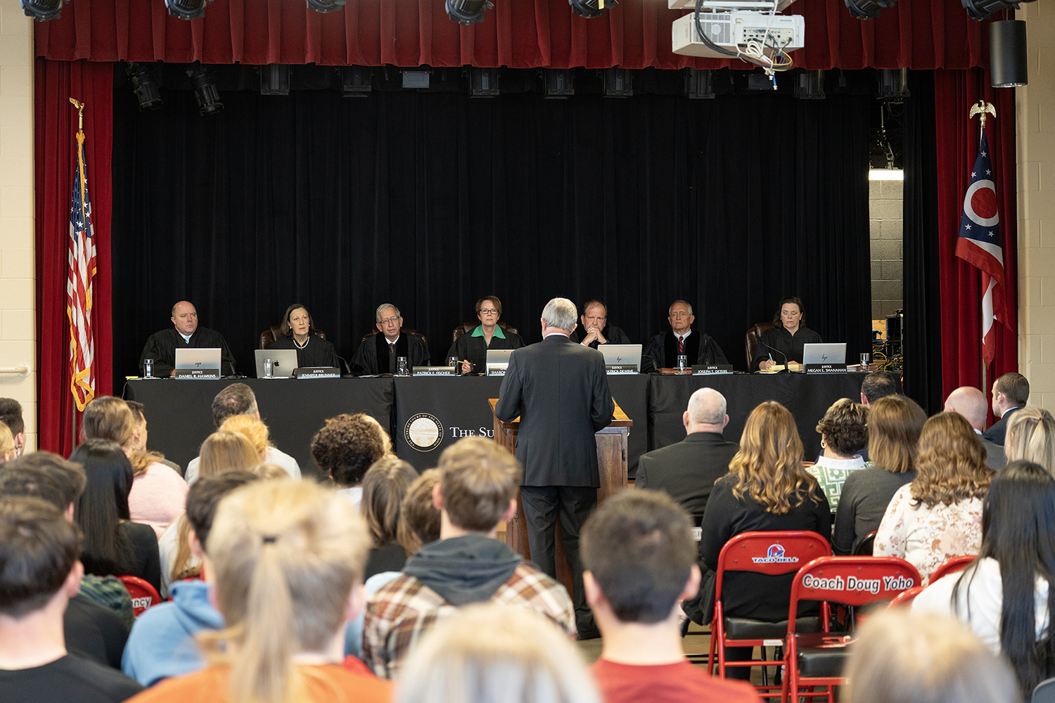 A man wearing a black suit stands at a wooden podium in front of a long table at which are seated seven men and women all wearing black judicial robes.