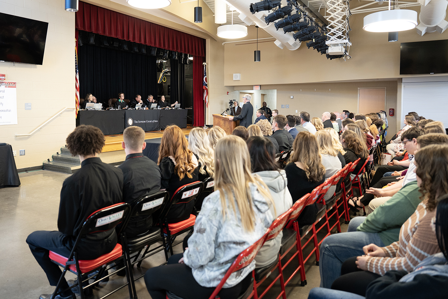 Rows of high school students watch a man in a black suit standing at a wooden podium addressing seven men and women all wearing black judicial robes seated at a long table.