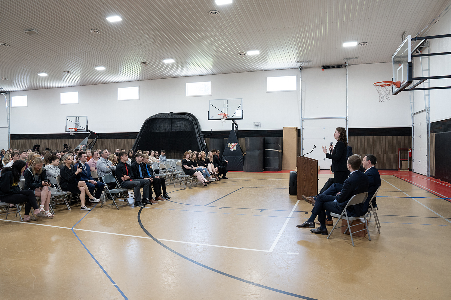 A woman wearing a black suit stands holding a microphone speaking to rows of high school students in a gymnasium.