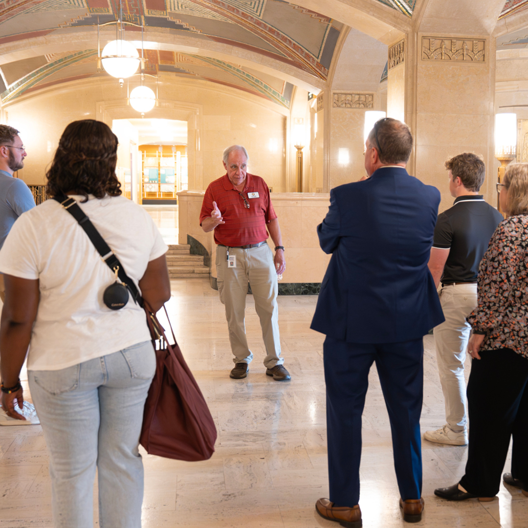 A group of men and women listen to a man in a red shirt speaking to them.