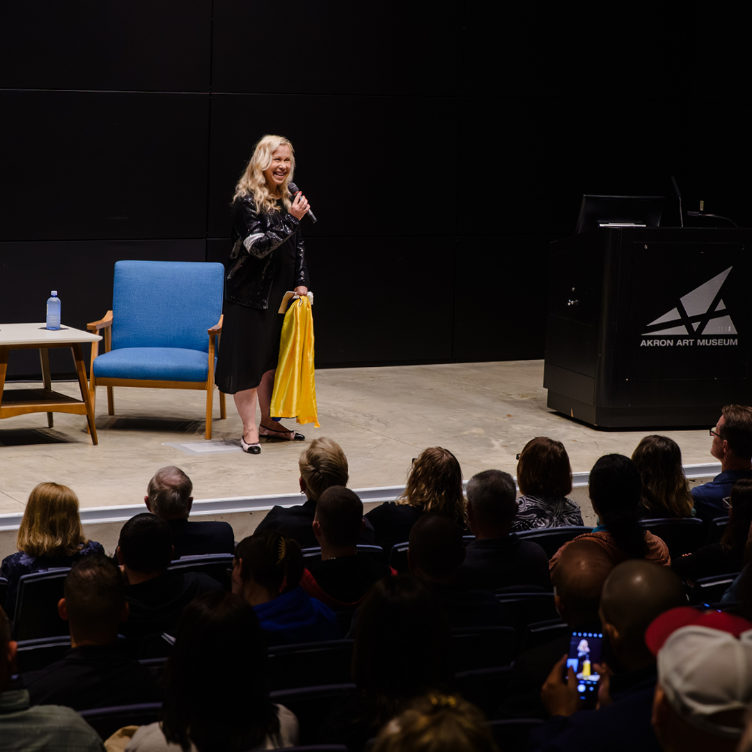 A woman wearing a black dress and holding a microphone in one hand and a yellow piece of fabric in the other hand speaks to an audience from a stage.