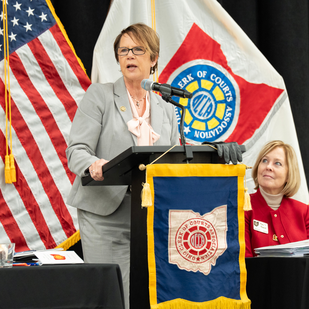 A woman speaking at a podiem with the US flag and Ohio flag in the background with a table of women listening to the speaker