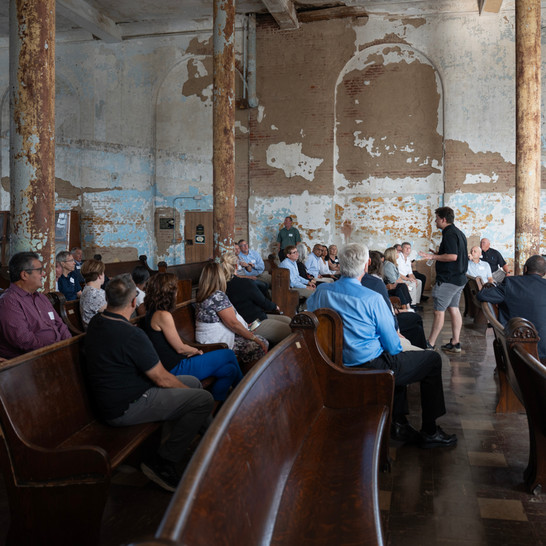 A group of men and women seated in wooden church pews in a very dilapidated room.