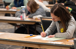 Men and women seated at long, wooden tables writing on papers with yellow pencils.