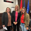 A woman wearing a brown polka dot blouse and black blazer, a woman wearing a bright pink polka dot blouse and black blazer, and a woman wearing jeans, a red shirt, and brown jacket standing side-by-side.