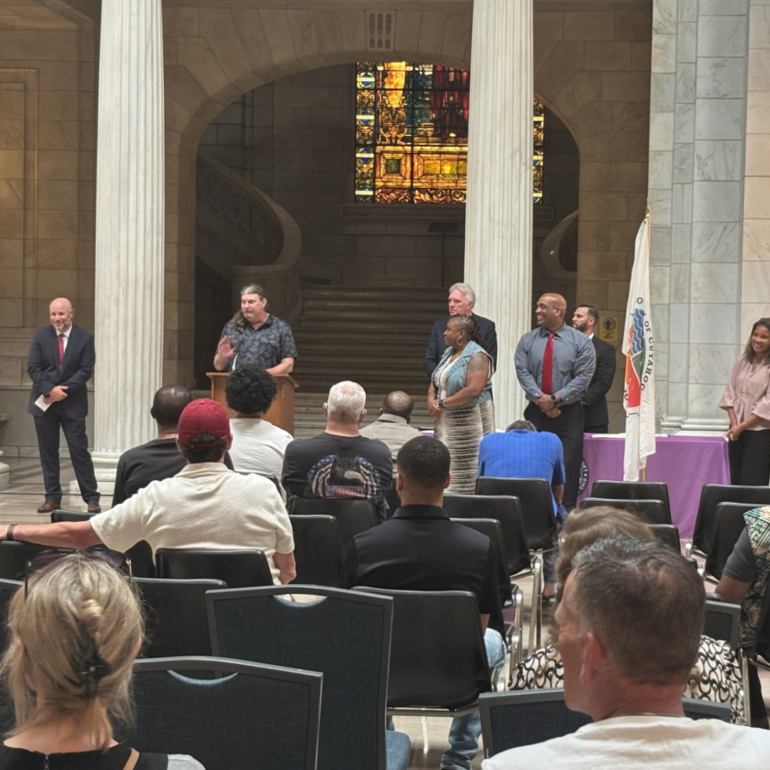 A man speaks to an audience from a wooden podium in a large room with marble columns and a marble staircase.