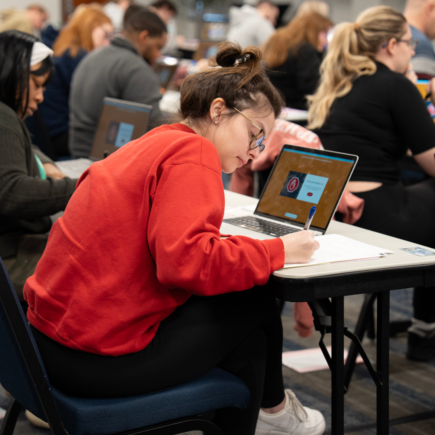 A student seated at a table writing on a piece of paper next to a laptop