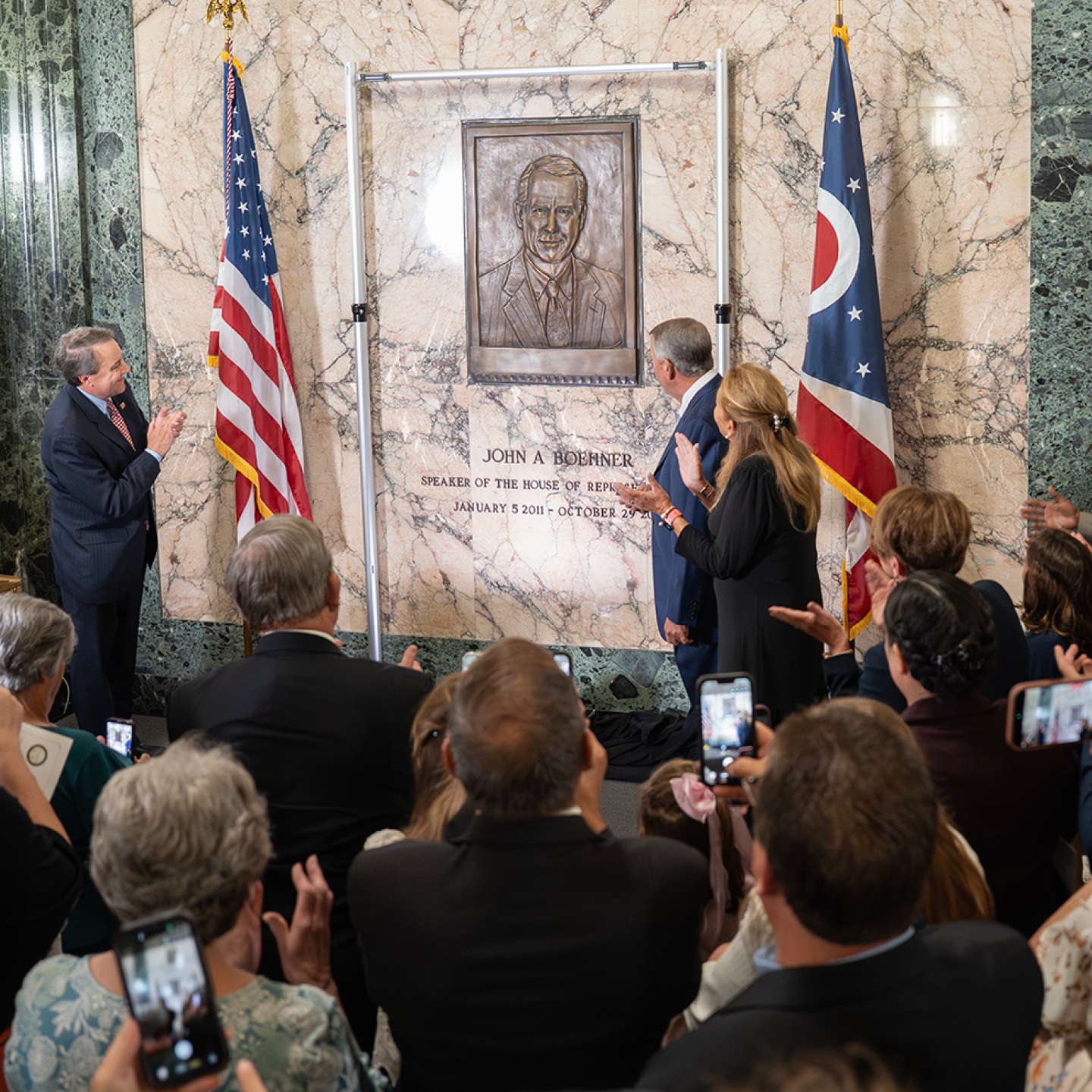 A crowd applauds a bronze plaque unveiling for John A. Boehner on a marble wall, flanked by U.S. and Ohio flags, conveying a celebratory mood.