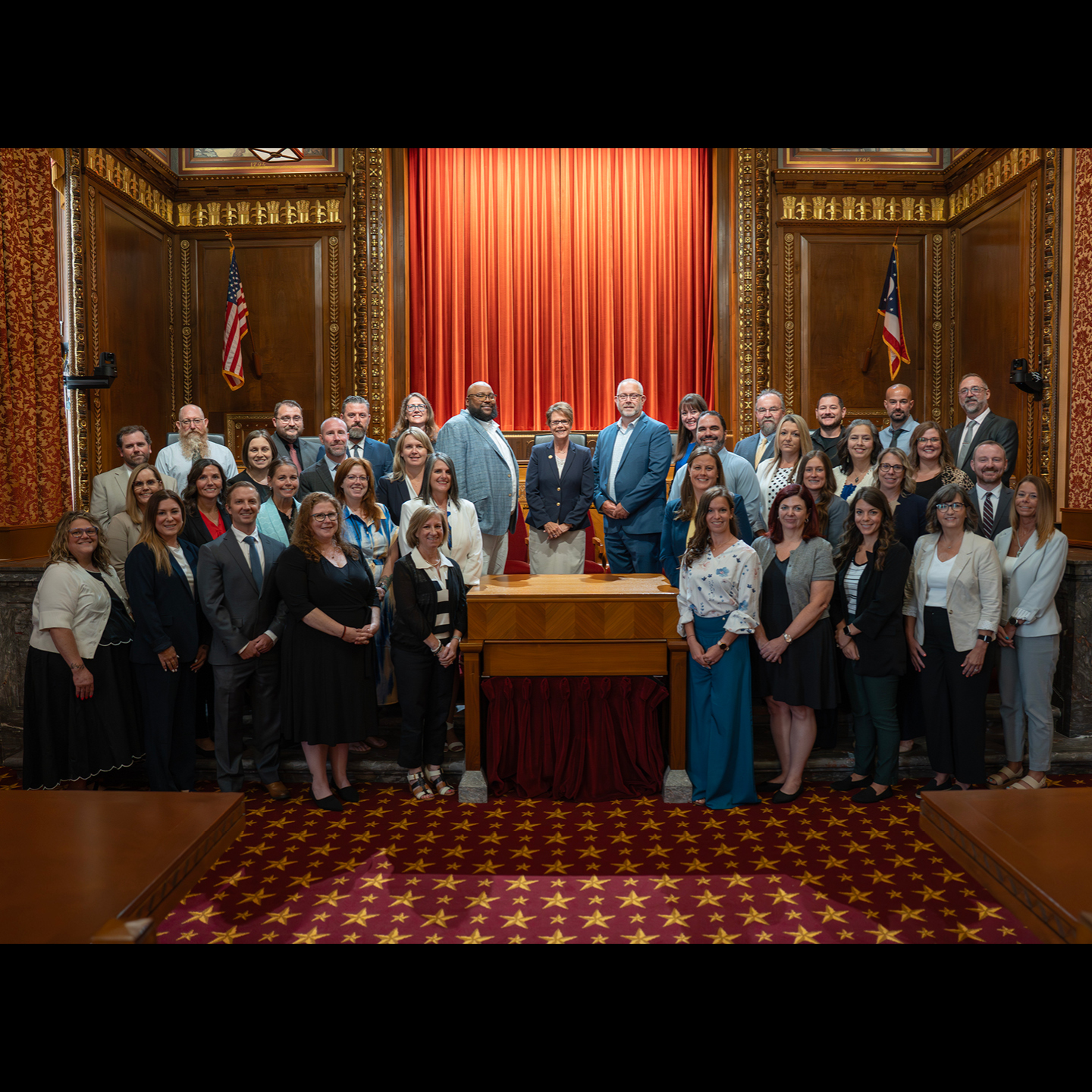 A large group of professionally dressed men and women pose for a group photo inside a grand, wood-paneled courtroom, standing in front of a wooden bench with a red curtain backdrop and the Ohio state flag.