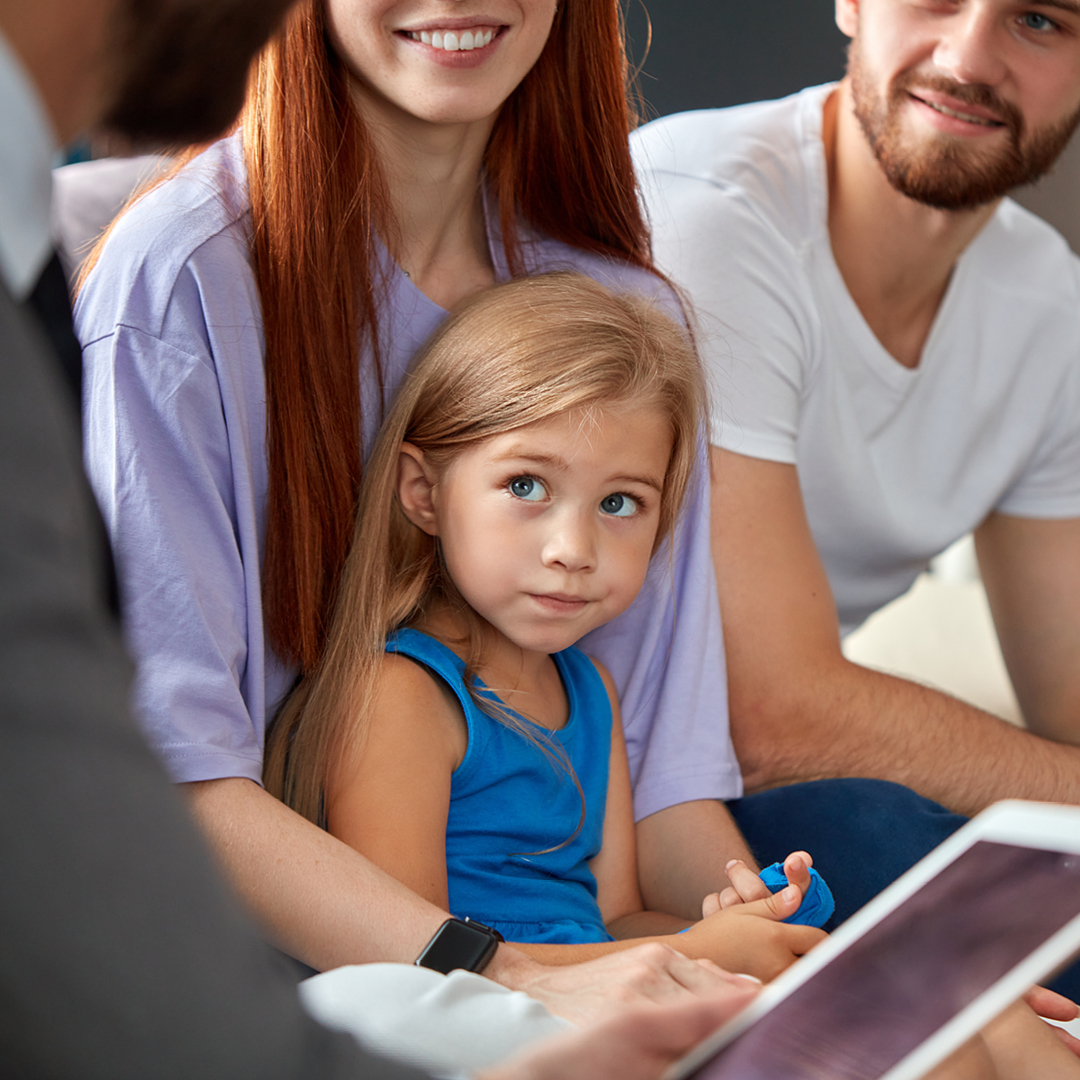 A woman with long, red hair and wearing a purple blouse holds a little girl. A man with a beard and white t-shirt sits next to them