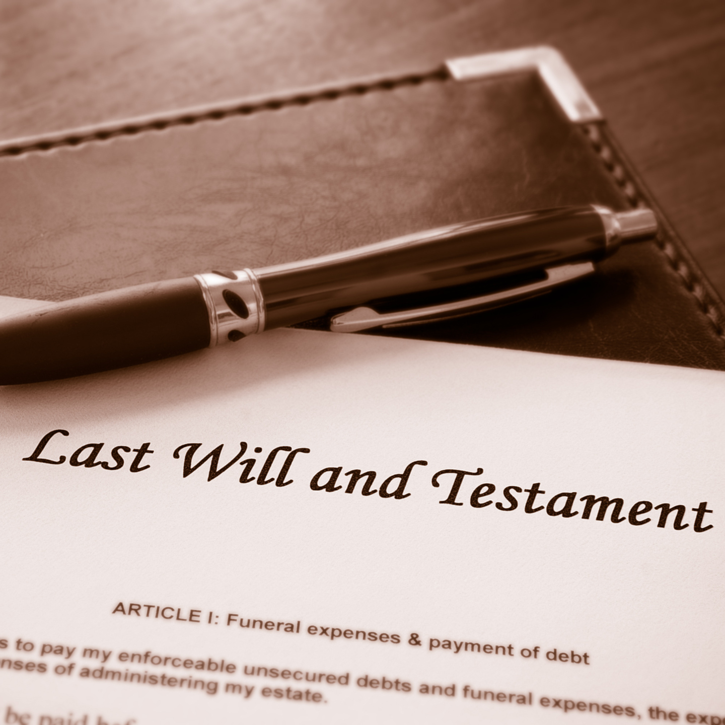 A sepia-toned, close-up photograph of a 'Last Will and Testament' document, with a pen resting on a leather folder in the background.