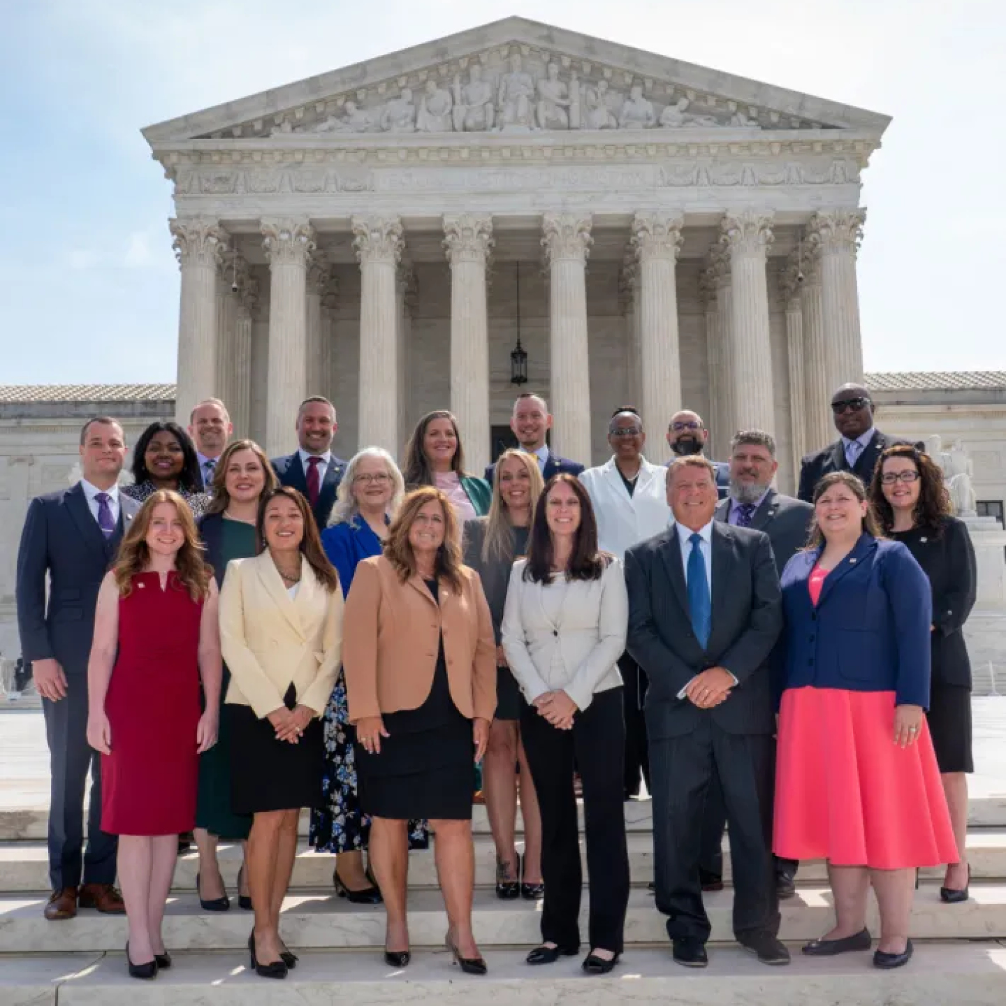 A group of professionally dressed individuals stands smiling on the steps of the Supreme Court Building in Washington, D.C.