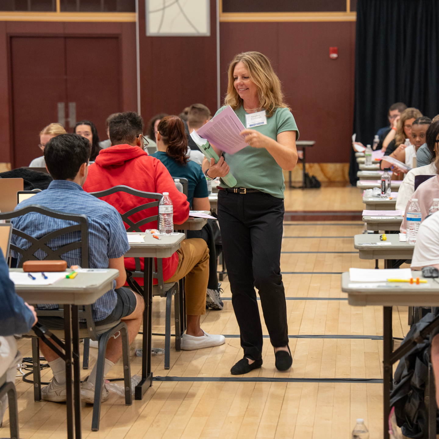 A woman in a green top and black pants walks through a classroom, holding papers and smiling, while students are seated at tables focused on their work.