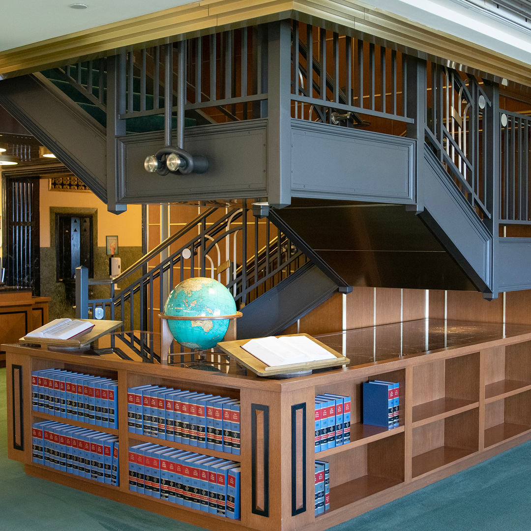A library with wooden shelves filled with books under a dark metal staircase. A globe and open books rest on the shelves.