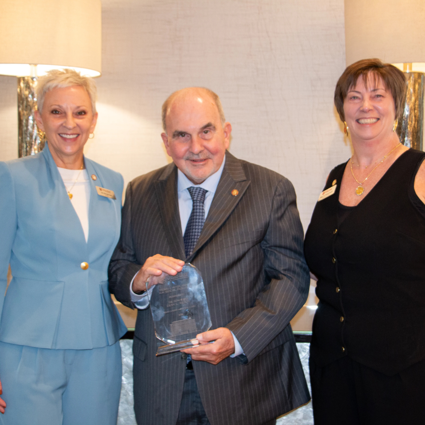A man in a pinstripe suit stands smiling between two women while holding a clear acrylic plaque award. The woman on the left has short, cropped gray hair and wears a light blue pantsuit with gold buttons over a white top. The woman on the right has shoulder-length brown hair with bangs and wears a sleeveless black top with gold buttons.