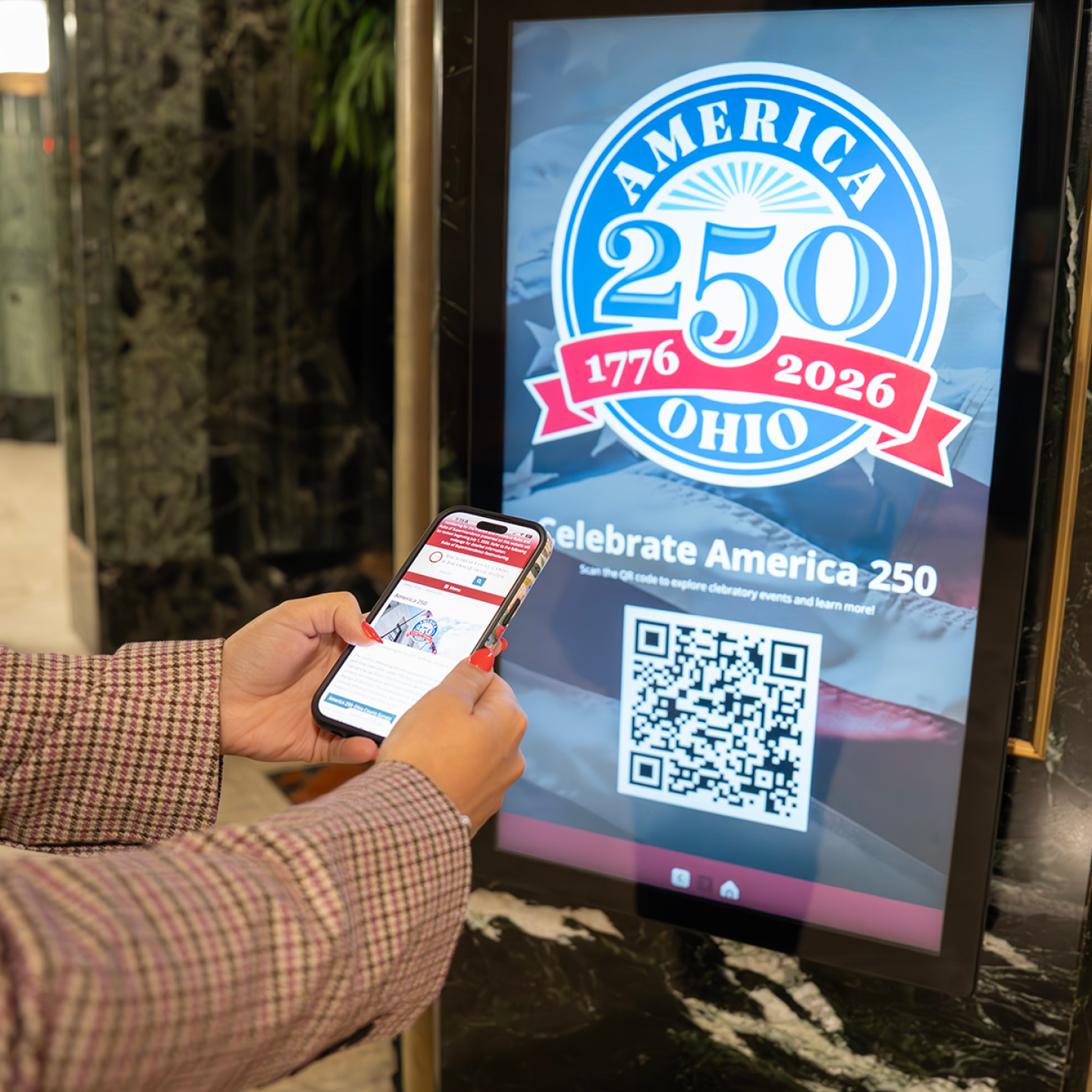A person uses a smartphone to interact with a digital display advertising the America 250 celebration in Ohio, featuring a prominent logo and QR code for event information.