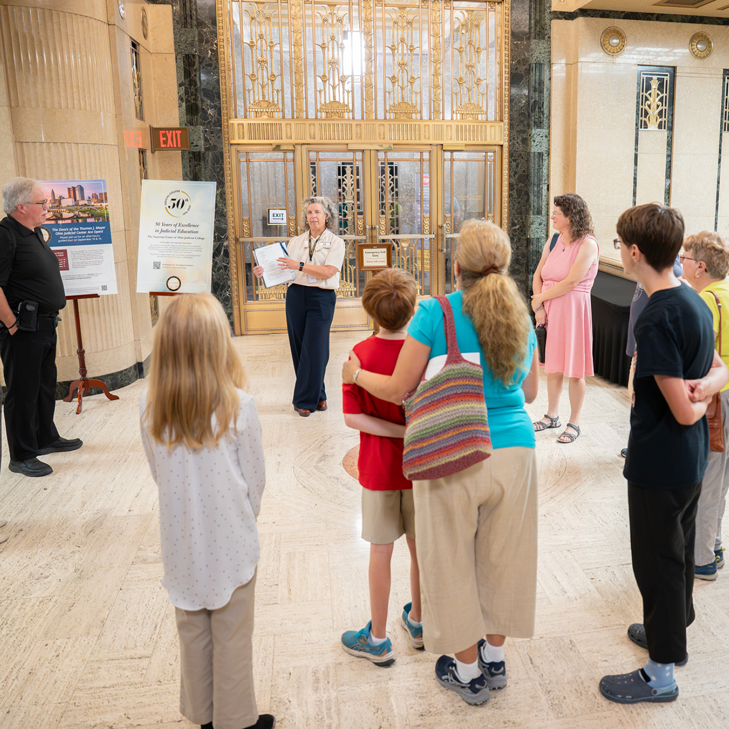 A group listens to a speaker in an ornate lobby with a gold-grilled elevator and informational displays.