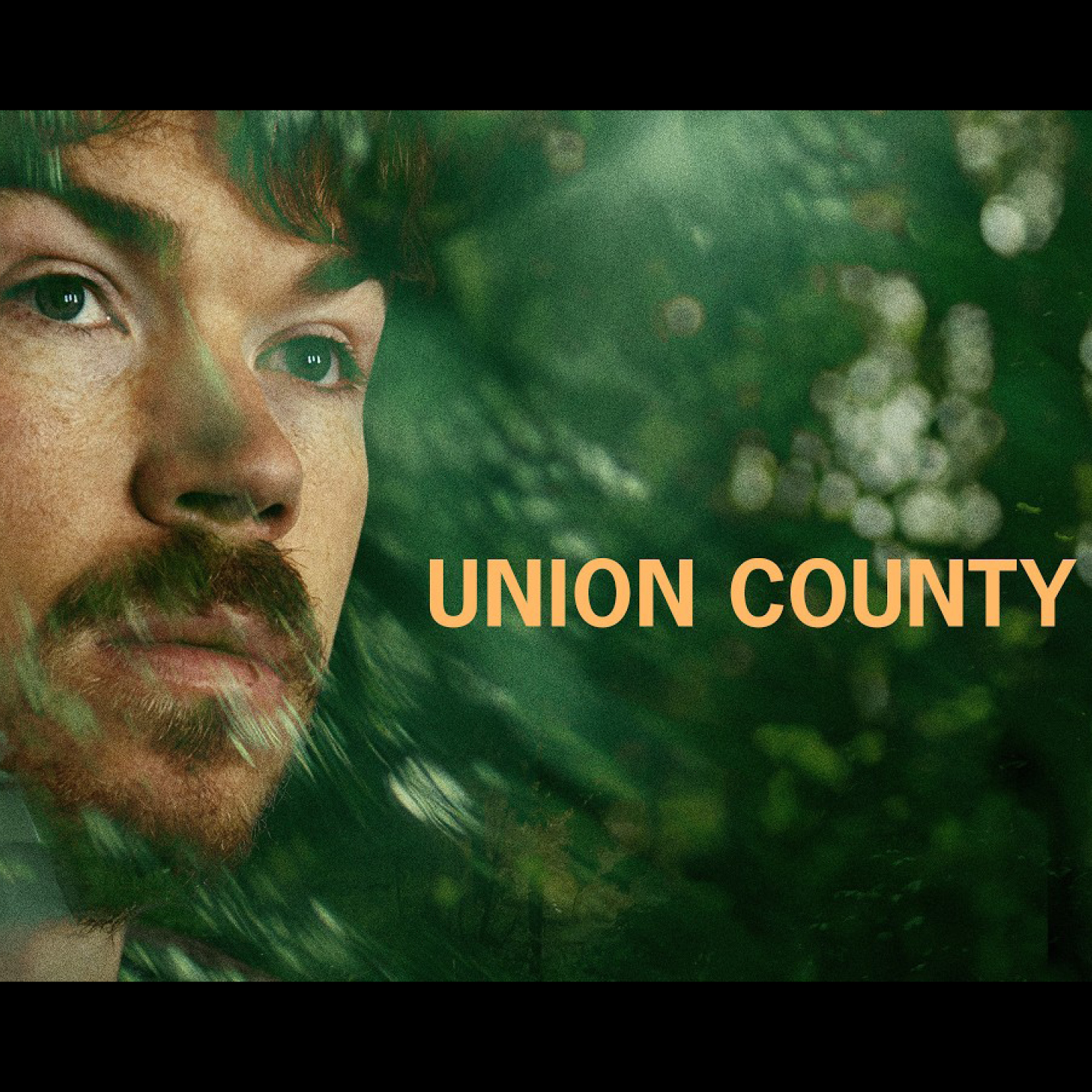 Close-up portrait of a bearded person against blurred green foliage, with the words “UNION COUNTY” in large yellow-orange letters.
