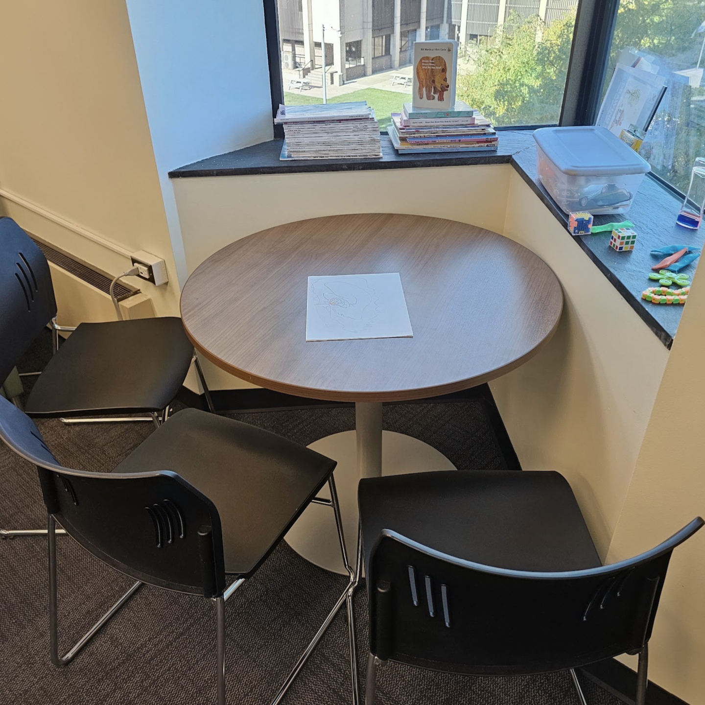 Small reading area with a round table, black chairs, books, toys, and a view of a courtyard through the windows.
