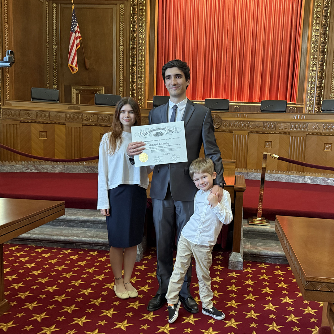A man in a suit holds a certificate, standing proudly with a woman and a child in a formal courtroom. Red curtains and an American flag are visible.