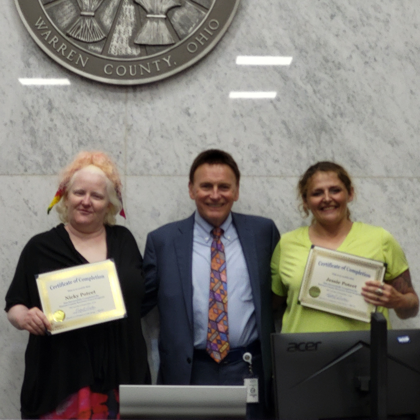 Three adults pose with framed certificates beneath a partial Warren County, Ohio seal.