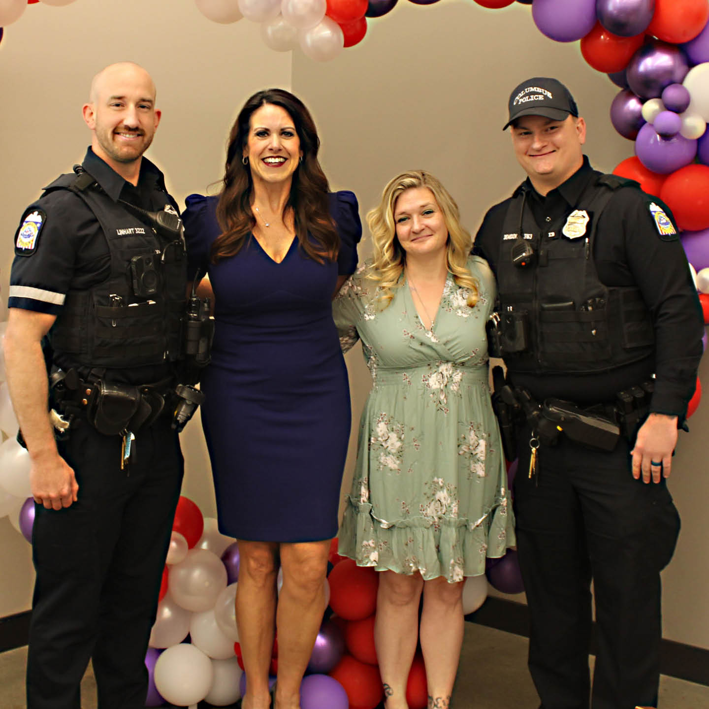 Four people stand smiling in front of a colorful balloon arch. Two are in police uniforms, and two women are wearing a blue dress and a floral dress.