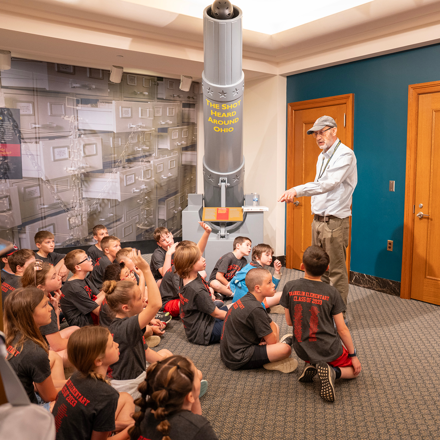 Children sit on the floor listening to an older man beside a tall exhibit labeled 'THE SHOT HEARD AROUND OHIO.'