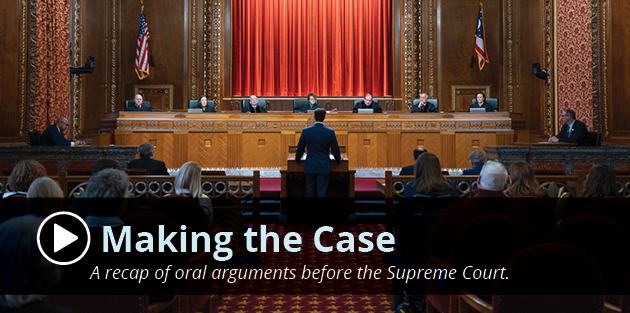 A man wearing a suit stands at a wooden podium facing seven men and women, all wearing black judicial robes, seated at a wooden bench in the courtroom of the Thomas J. Moyer Ohio Judicial Center.