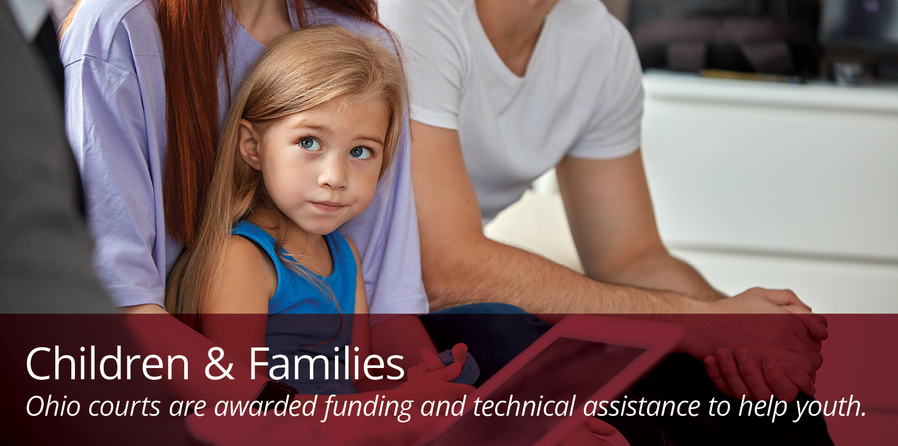 A woman with long, red hair and wearing a purple blouse holds a little girl. A man with a beard and white t-shirt sits next to them