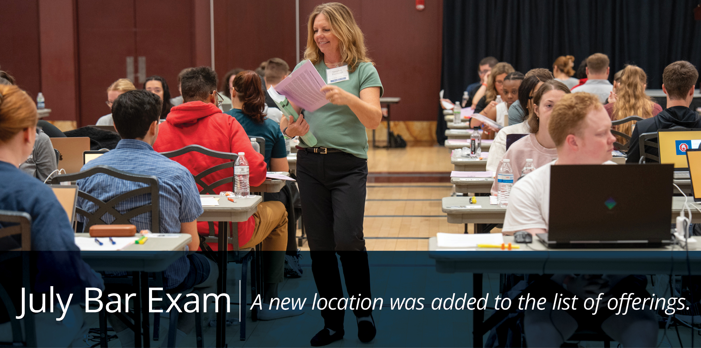 A woman in a green top walks through a room of students taking the July Bar Exam, holding papers and smiling. Students are at tables with laptops and water bottles.