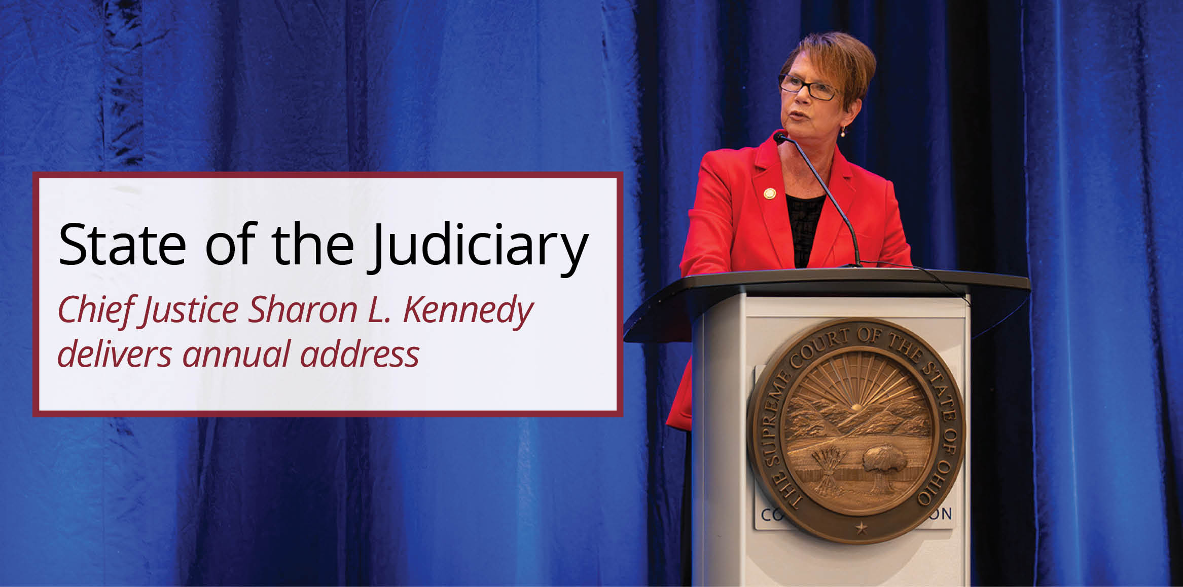 A woman with short brown hair and glasses, wearing a bright red blazer over a black top, speaking at a podium.