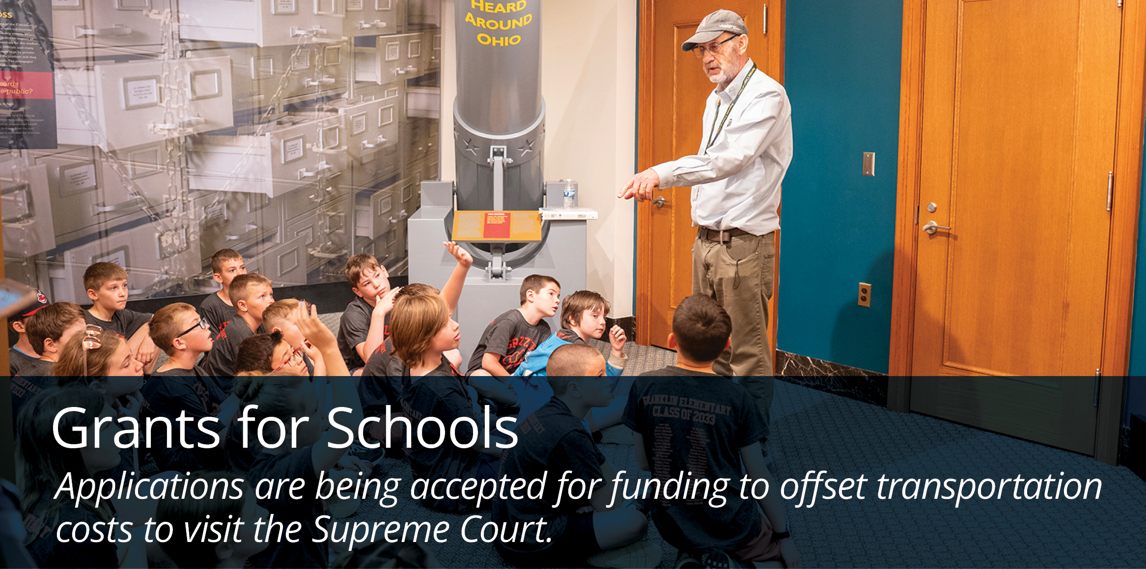 Schoolchildren listen and raise their hands during a presentation in an exhibit space, with a banner announcing grants to offset transportation costs for visits to the Supreme Court.