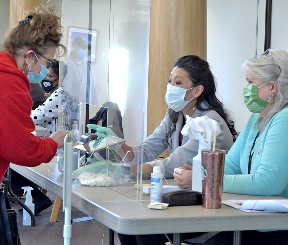 Image of two women wearing masks sitting behind a plexiglass barrier assisting a voter