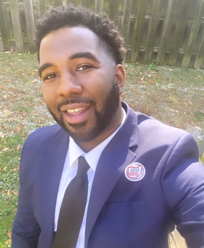 Image of a man wearing a blue suit with an Ohio voting sticker on the lapel standing in front of a wooden fence taking a selfie