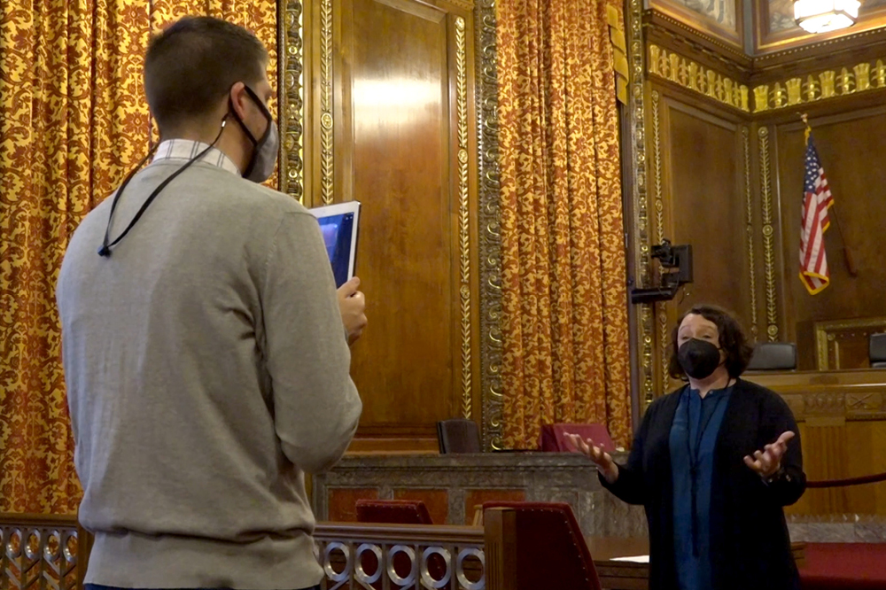 Image of a man holding up a tablet in front of a woman speaking in the courtroom of the Thomas J. Moyer Ohio Judicial Center