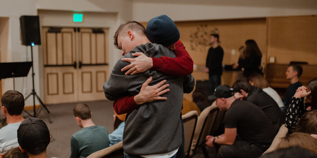 Image of two young men embracing each other amongst a group of men and women sitting in chairs