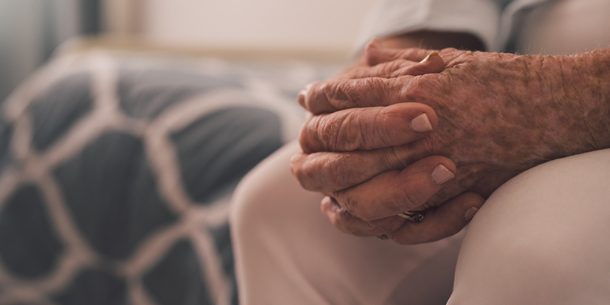 Image of an elderly woman sitting on the edge of a bed with her hands clasped