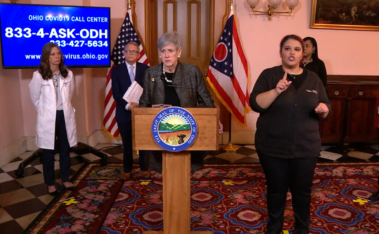 Image of a woman speaking from behind a podium. Beside her is a woman translating in American Sign Language. Behind her stand a woman wearing a white doctor's coat and a man wearing a suit