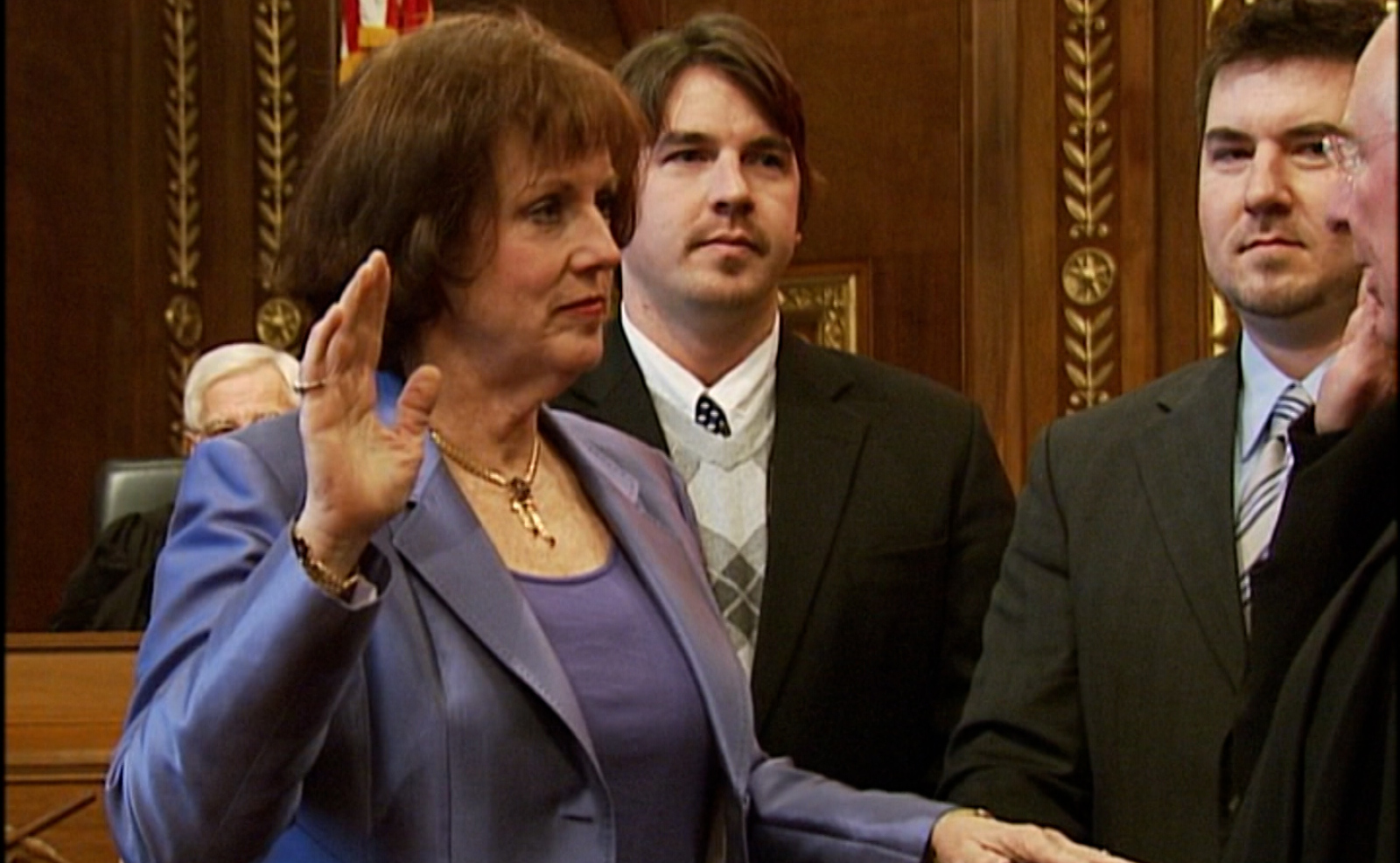 Image of a woman wearing a blue suit holding her right hand up. Two me in suits stand behind her