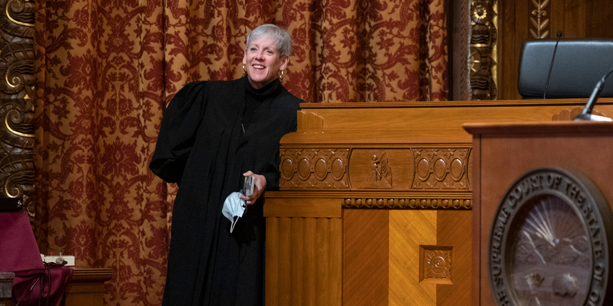 Image of Ohio Supreme Court Chief Justice Maureen O'Connor in her black judicial robe smiling and leaning against the bench in the courtroom of the Thomas J. Moyer Ohio Judicial Center