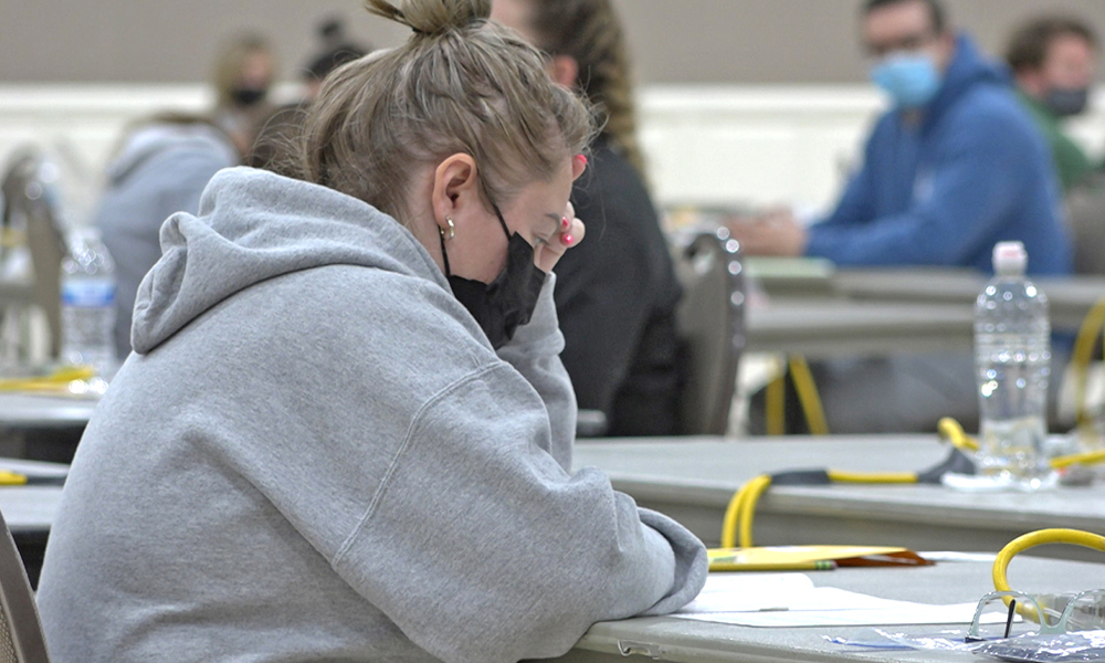 Image of a woman wearing a grey hoodie and a black mask sits at a table taking an exam
