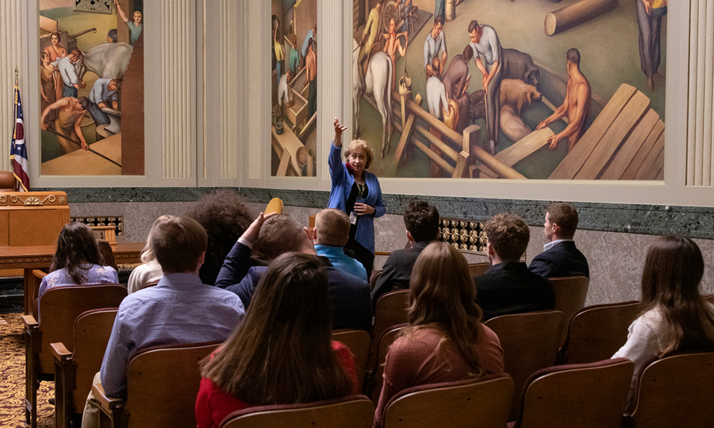 Image of a woman with her hand raised in the air as she speaks to a group of students while standing in front of a mural in the North Hearing Room of the Thomas J. Moyer Ohio Judicial Center