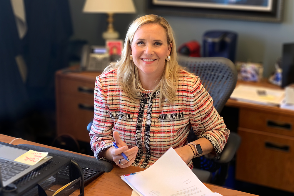 Image of a woman with long blond hair sitting at a desk and smiling