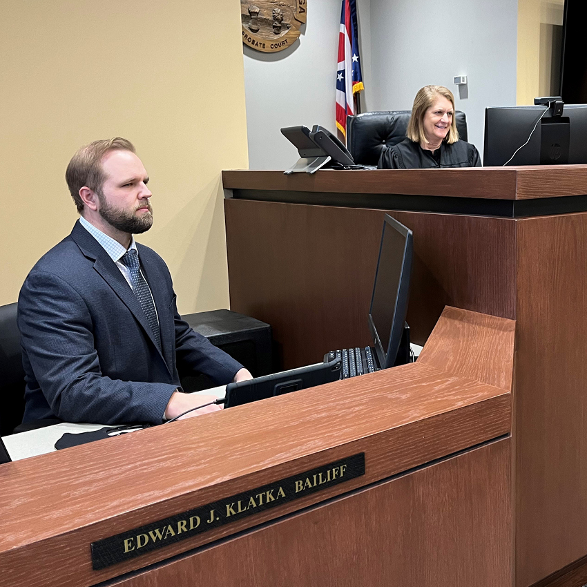 Image of a man wearing a blue suit sitting at a desk next to a judge's bench