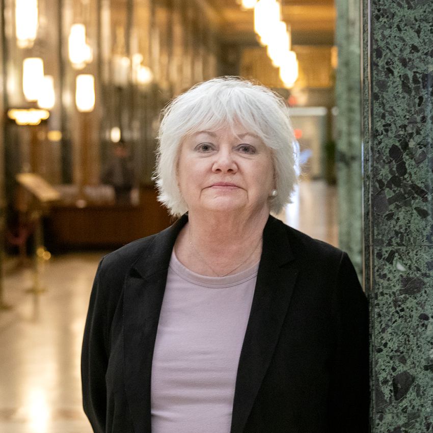 Image of a woman wearing a grey top and black blazer standing next to a green marble wall