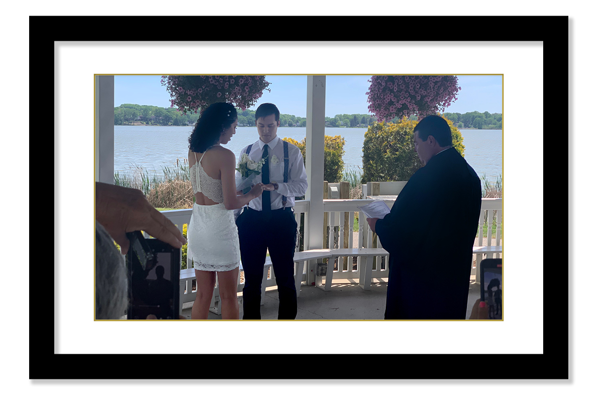 Image of a woman wearing a white, lacy dress placing a ring on the finger of a man dressed in dark pants, white dress shirt, and suspenders. The couple is standing in a gazebo by a lake. A man wearing a black judicial robe and holding a paper stands in front of the couple.