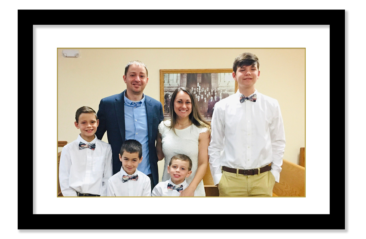 A man wearing a blue dress shirt and blue suit jacket stands next to a woman wearing a white, lacy dress. Four boys, all wearing matching outfits stand alongside the couple.