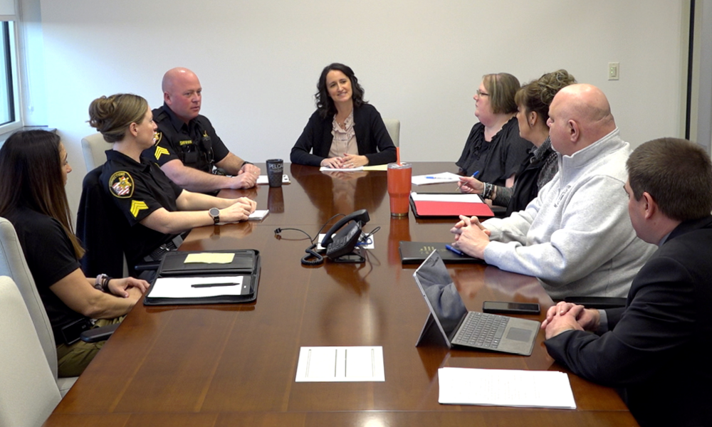 Image of several men and women, some wearing law enforcement uniforms, others wearing dress clothes, seated at a long, rectangular table.