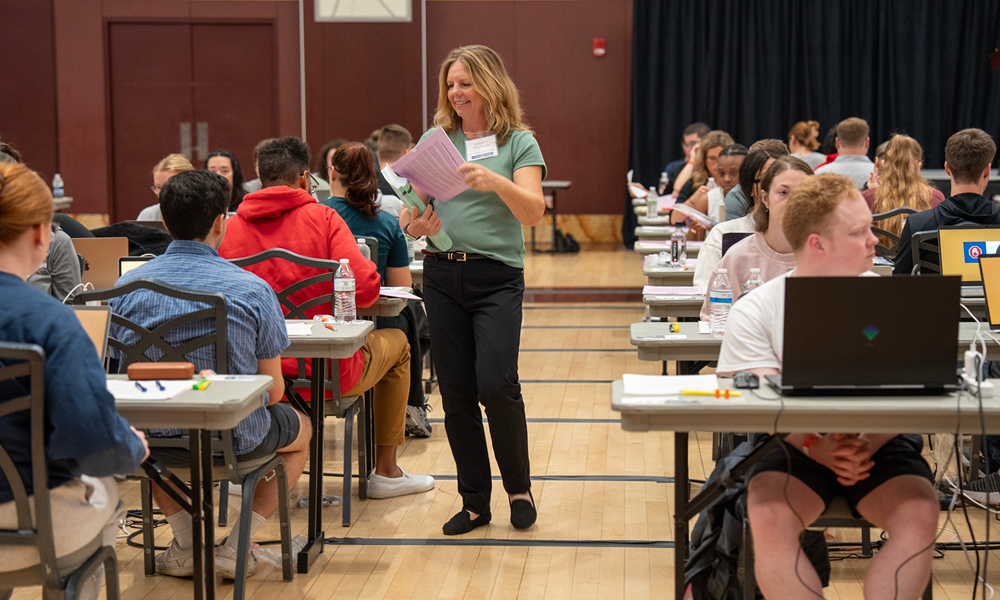 A woman wearing black pants and a green, short-sleeve shirt passes out papers to a roomful of men and women seated at long tables.